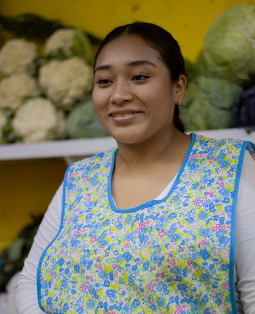 Free Smiling woman wearing apron stands in front of fresh vegetables at market stall. Stock Photo