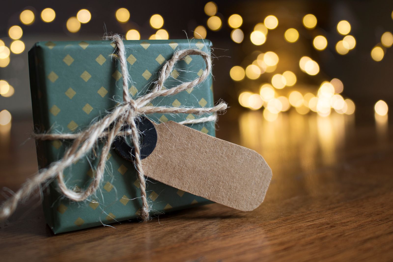 A wrapped gift box with twine beside festive bokeh lights indoors.
