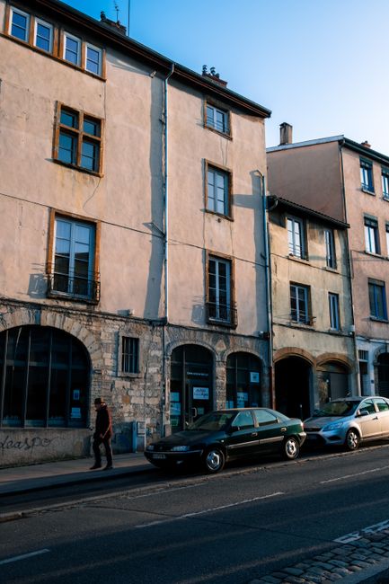 Street view of historic city architecture with sunlit facades and parked cars on a clear day.