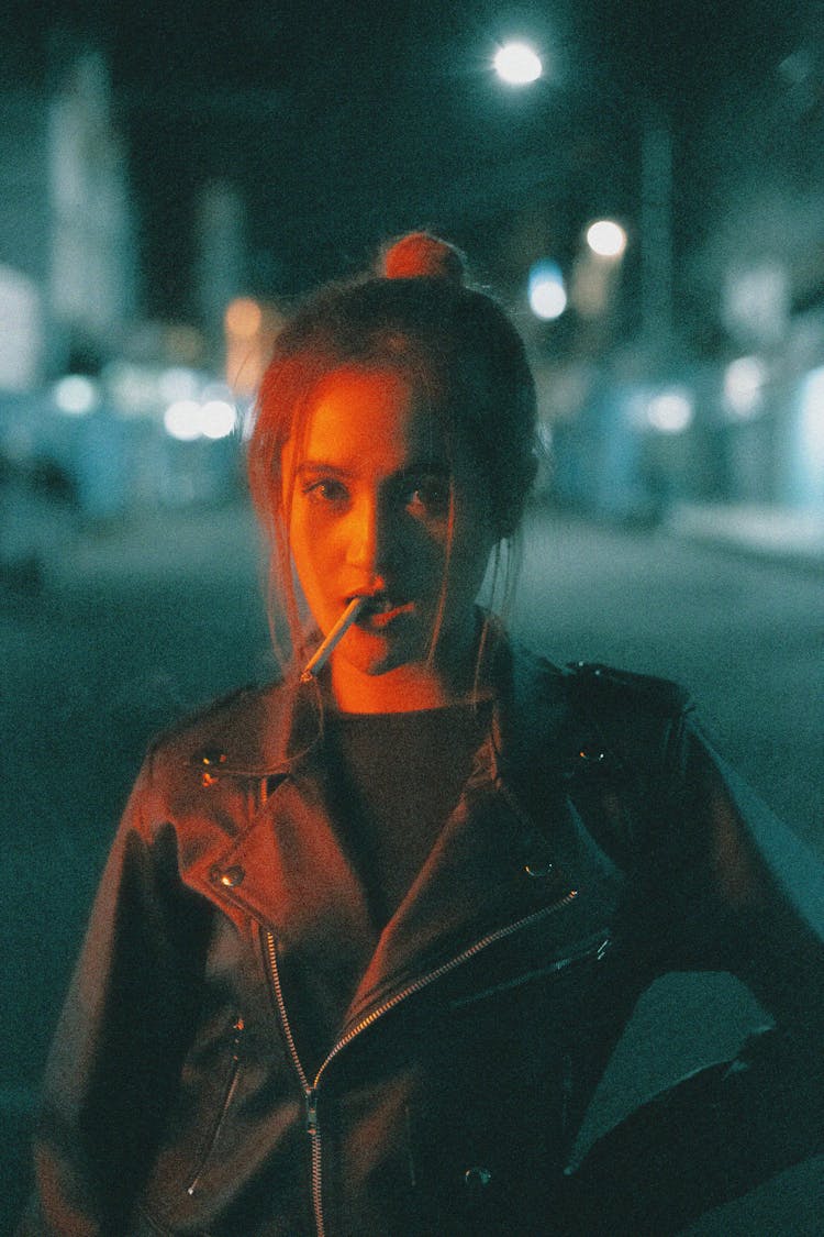 Young Woman In A Leather Jacket Standing On A Street In City At Night And Smoking 