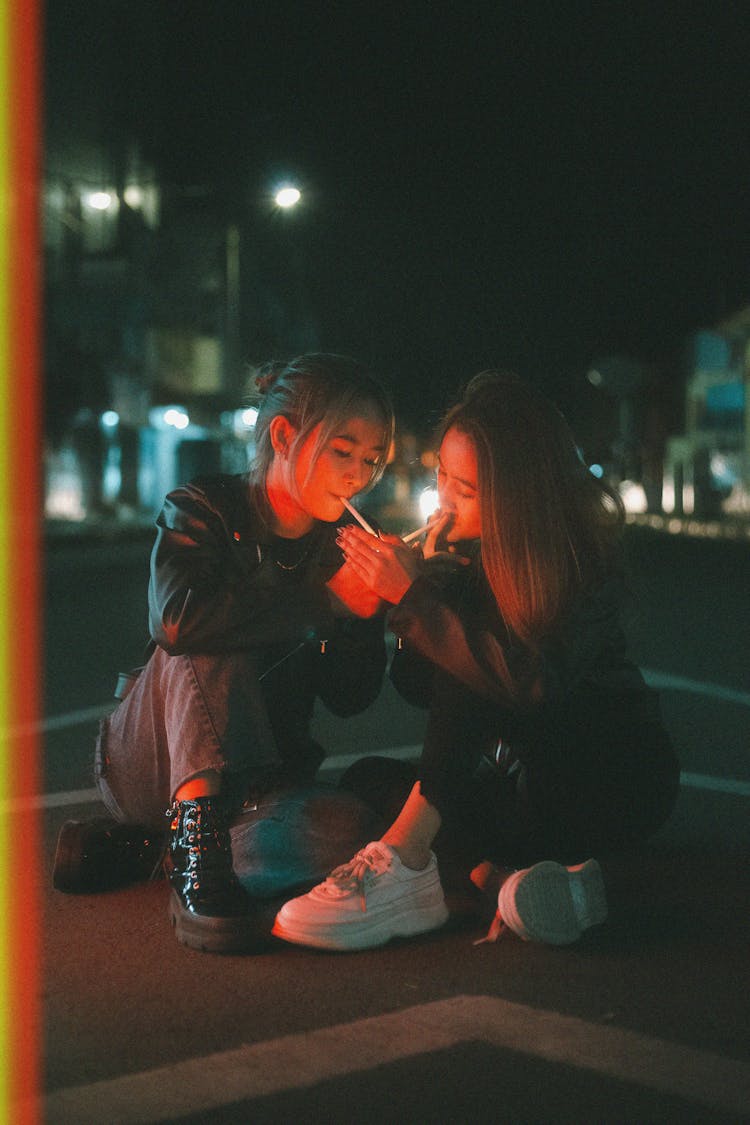 Two Young Women In Leather Jackets Sitting On A Street At Night And Smoking 