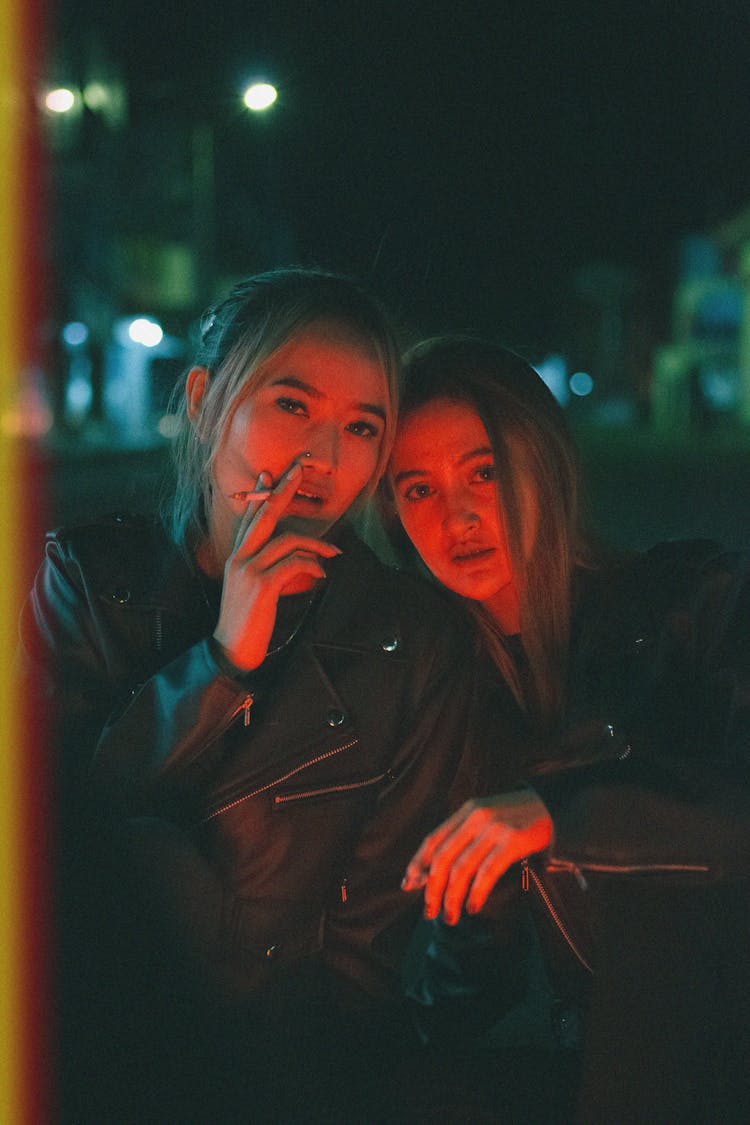 Two Young Women In Leather Jackets Sitting On A Street At Night And Smoking 