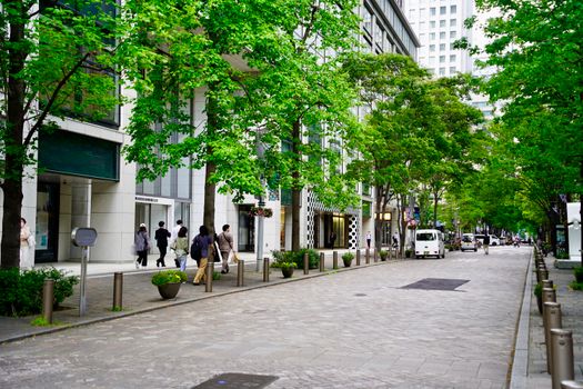 A lively urban street scene with lush green trees and people walking on a paved road in a modern cityscape.