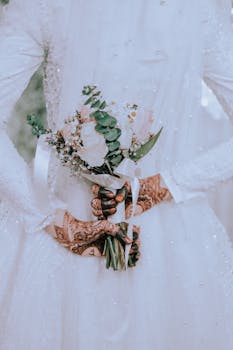 Close-up of a bride in a white gown holding a floral bouquet, hands adorned with henna.