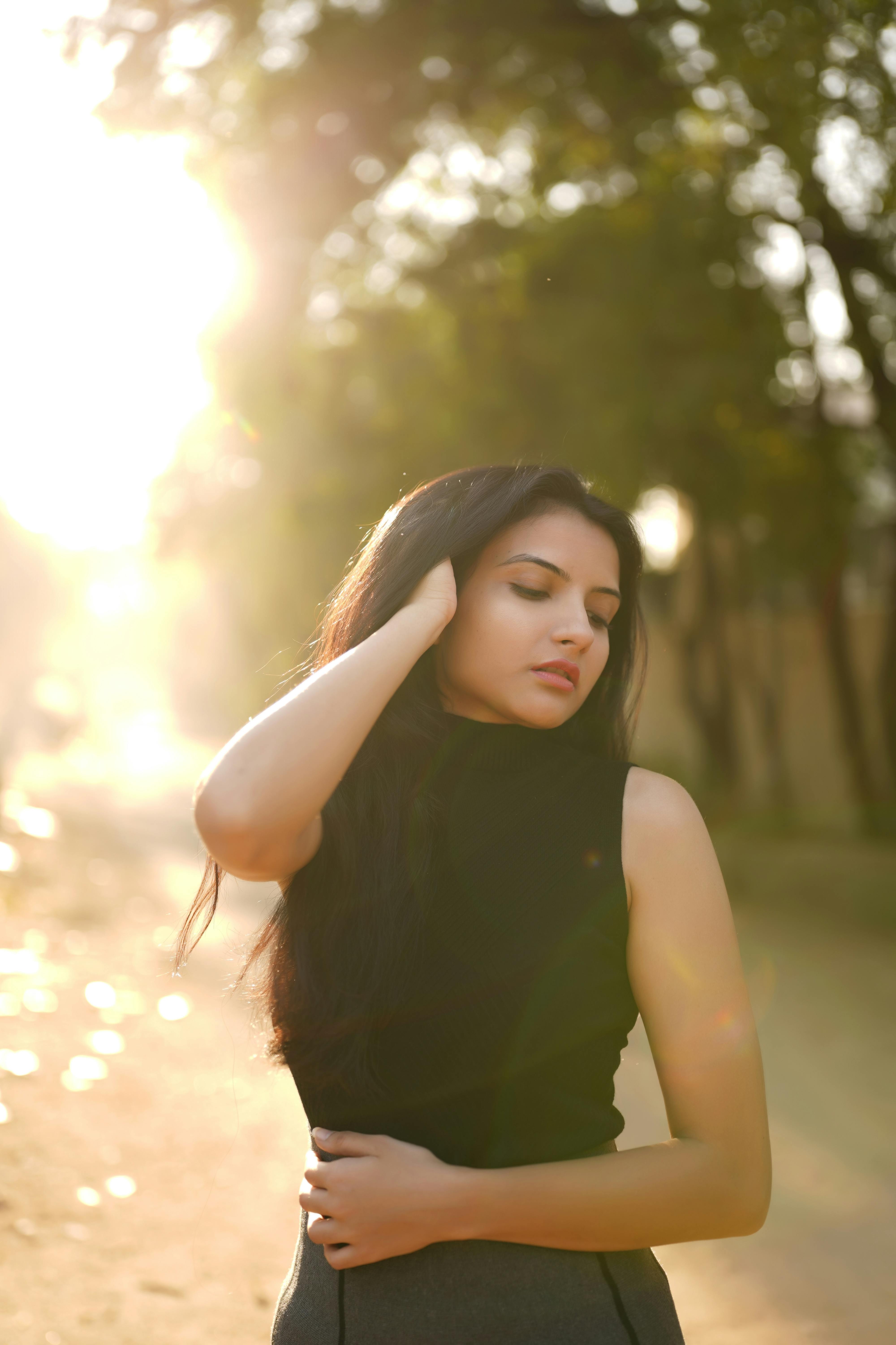 Elegant woman in black fashion, posing outdoors with sunlight and trees in the background.