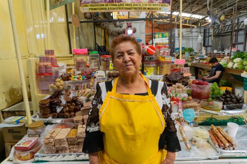 Free A cheerful woman in a yellow apron stands at her colorful market stall filled with local goods. Stock Photo