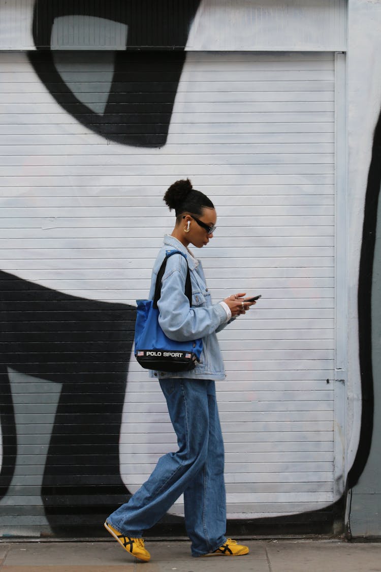 Side View Of A Woman Walking With A Phone By A Wall With A Mural