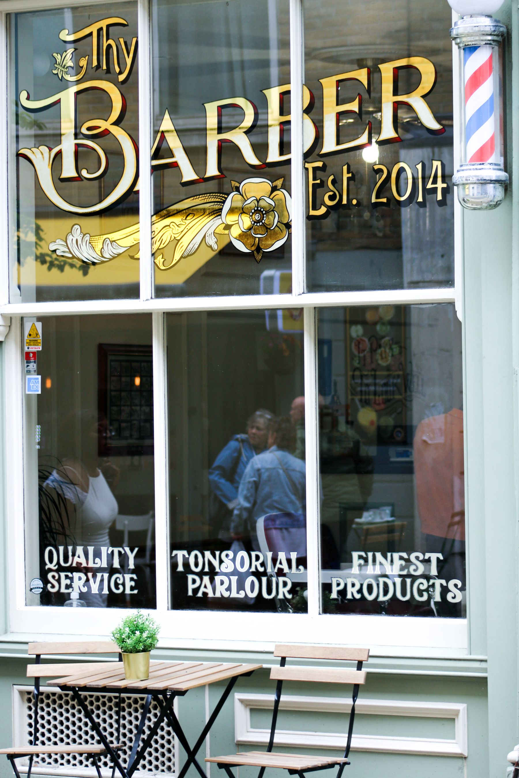 Real London barber shopfront with a barber pole, lettering on the window and an outdoor table and chairs