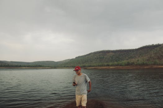 A man stands by a serene lake surrounded by hills, checking his phone under a cloudy sky.
