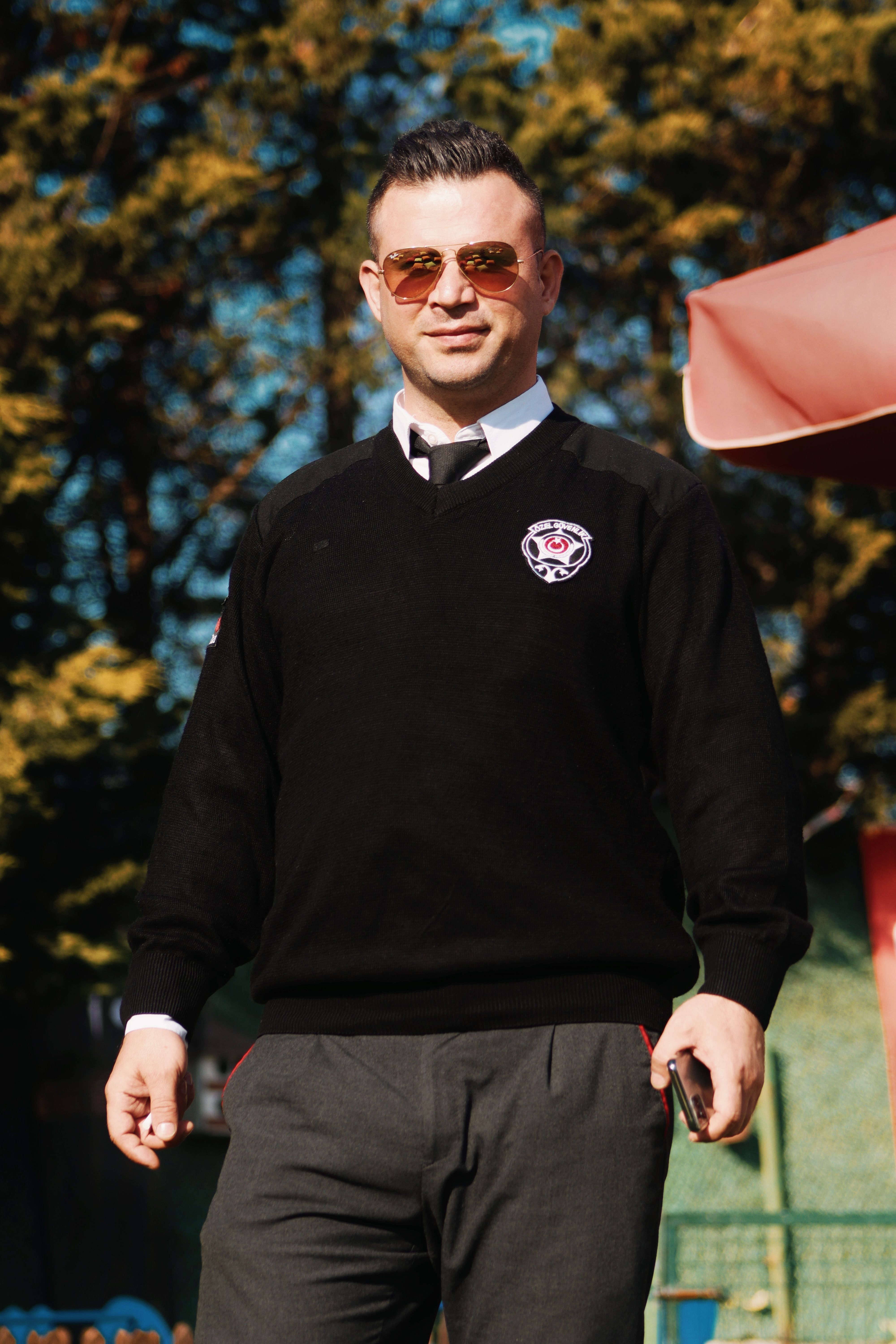 Stylish man wearing sunglasses and black sweater standing confidently in a park on a sunny day.