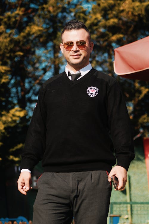 Photo of a Man Wearing a Black Sweater with a Logo Standing in a Park