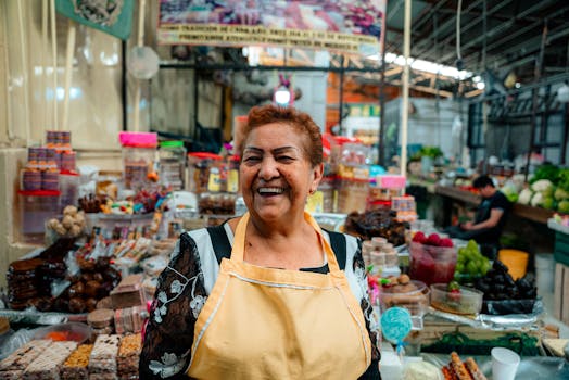 Smiling woman vendor in a bustling indoor market surrounded by colorful produce and treats.