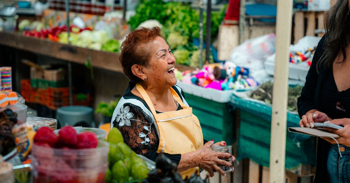 A traveler sitting on a wooden bench talking to an elderly local woman in a colorful market, both laughing