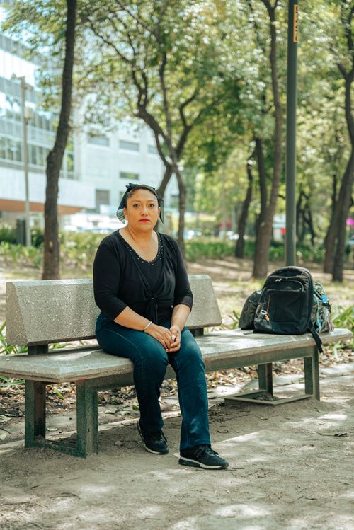 Free A woman sitting on a bench in a city park with a backpack beside her on a sunny day. Stock Photo