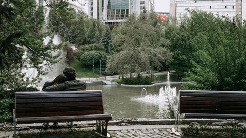A couple relaxes on a bench overlooking a peaceful fountain and greenery in a city park setting.