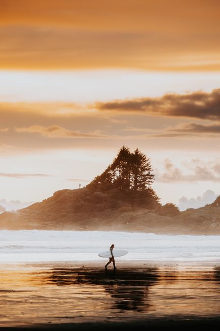 A lone surfer walks along the beach at sunset with a stunning island backdrop, capturing the serene coastal atmosphere.