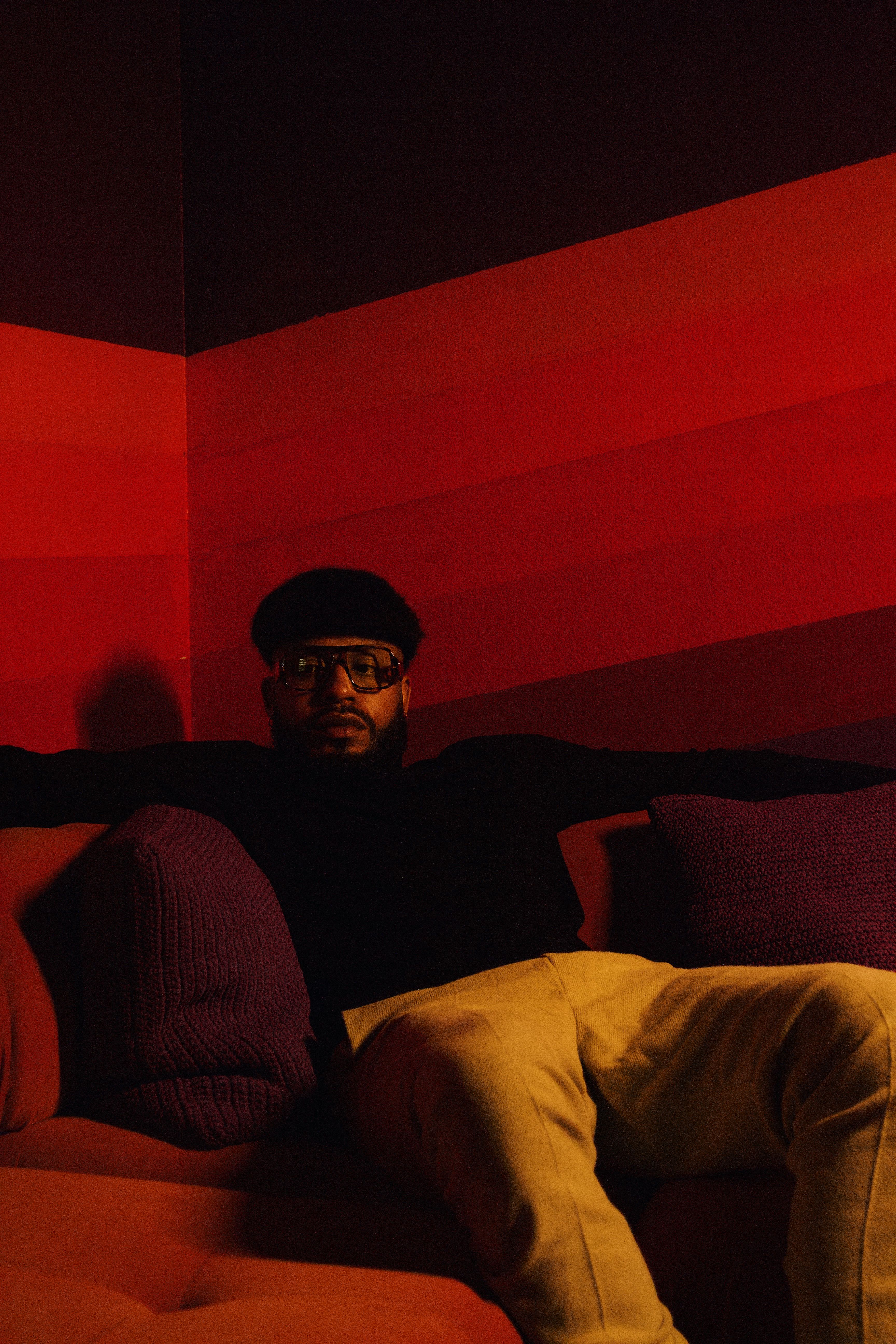 Man with eyeglasses and beard sitting on a sofa in a warmly lit room.