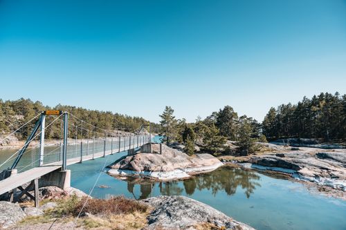 Free Wooden footbridge crossing a serene river surrounded by lush forest and rock formations. Stock Photo