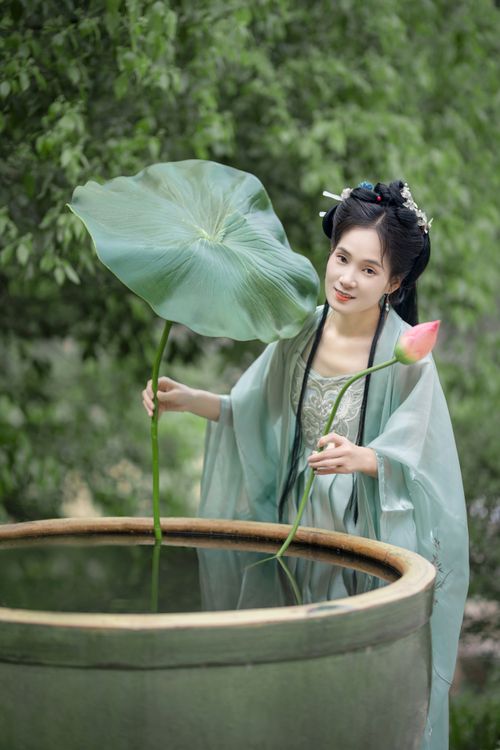 Free A woman in traditional clothing stands by a pond with lotus leaves, capturing a serene moment in a garden. Stock Photo