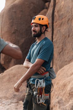 Man with climbing gear and helmet standing by rocky landscape, ready for adventure.