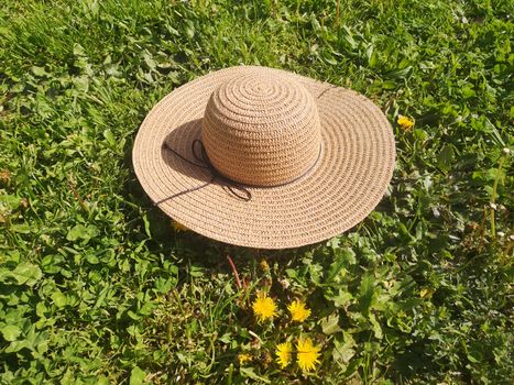 A straw hat sits on a vibrant green grass field, adorned with dandelions, under the summer sun.
