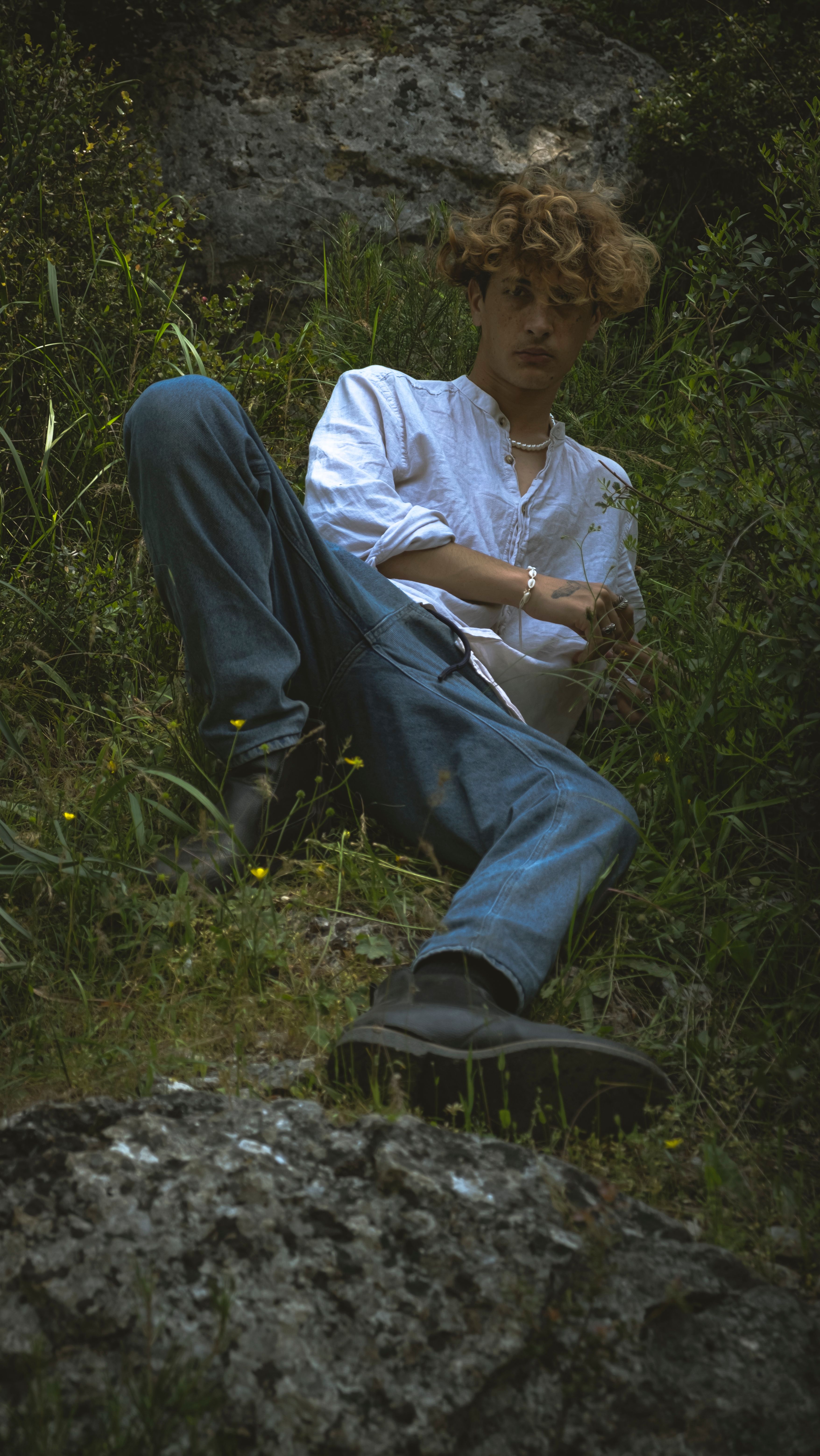 A man in a white shirt and jeans poses casually in a grassy outdoor setting.