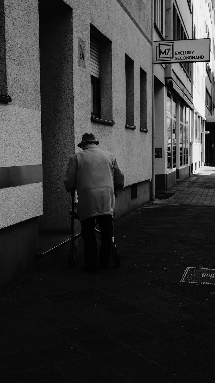 Elderly Man In Coat And Hat Walking Near Building In Black And White