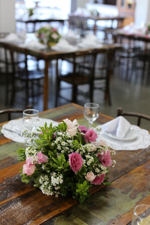 Pink And White Flower Centerpiece Beside Wine Glasses And Plates On Brown Table