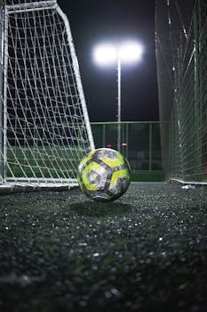 A football resting near the goal post under bright floodlights on a pitch at night.