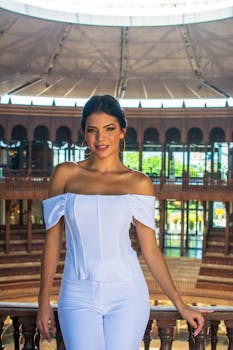 Elegant woman in white attire posing indoors with natural light and architectural backdrop.