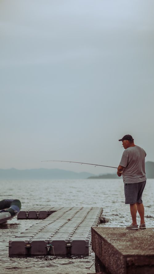 Free A man enjoys fishing on an overcast day by a tranquil lake from a pier. Stock Photo