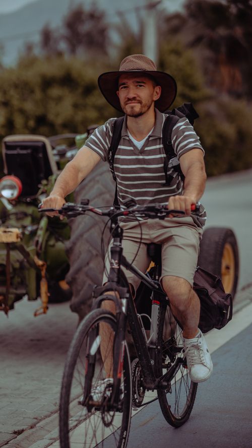 Free Man on bicycle with a backpack, wearing a hat, cycling past a tractor on a rural road. Stock Photo