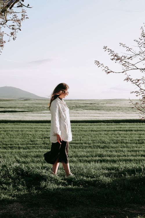 Free Woman in a white shirt walking in a green field under a clear sky, embodying tranquility and nature's beauty. Stock Photo