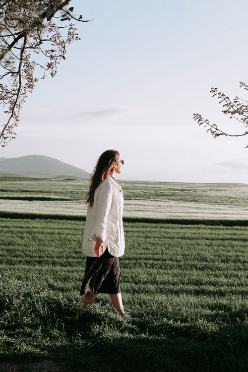 Free A woman with long hair walks through a peaceful green field wearing a white jacket. Scenic rural setting. Stock Photo
