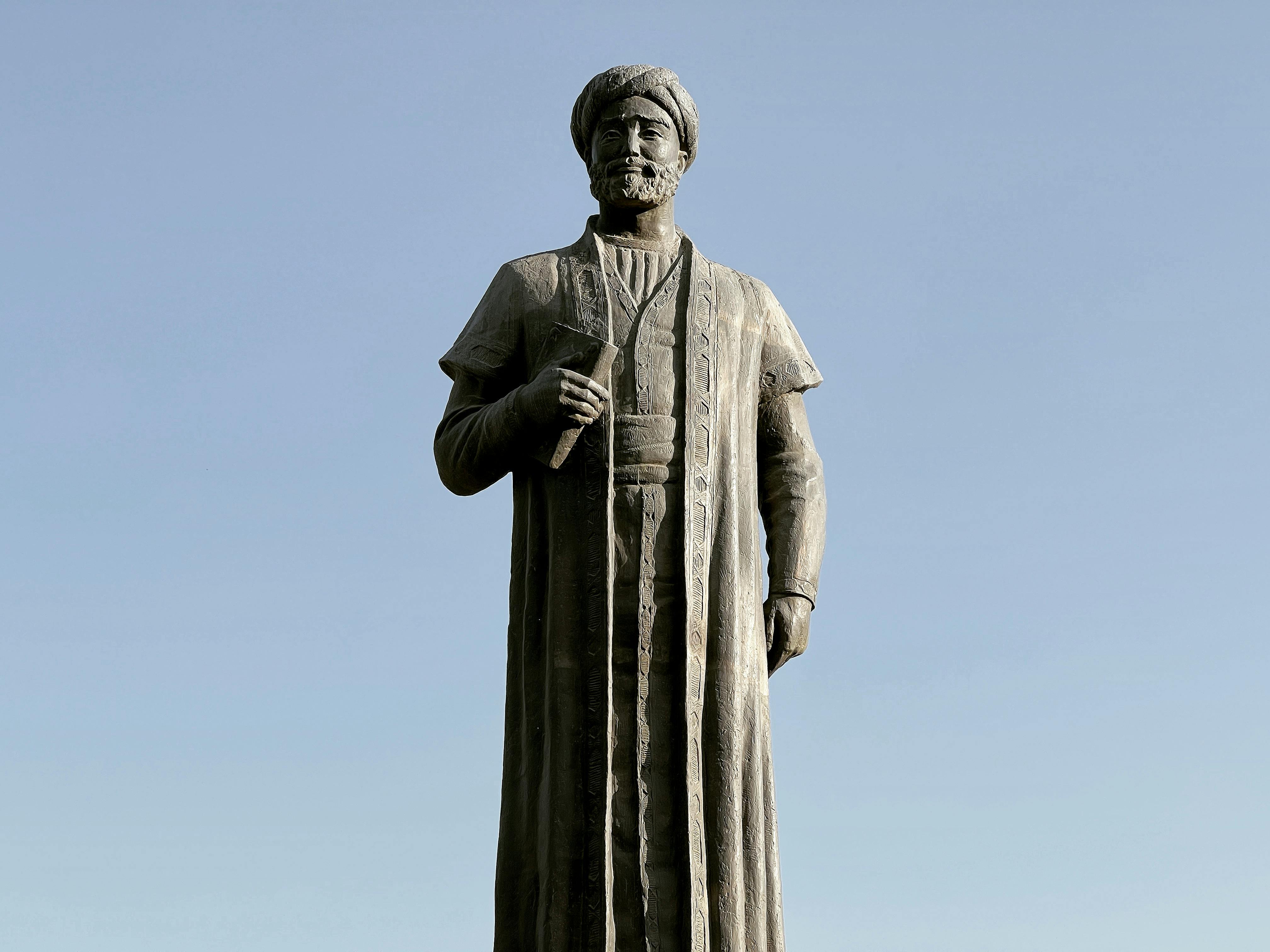 A stone statue of a historic figure holding a book stands under a clear blue sky.