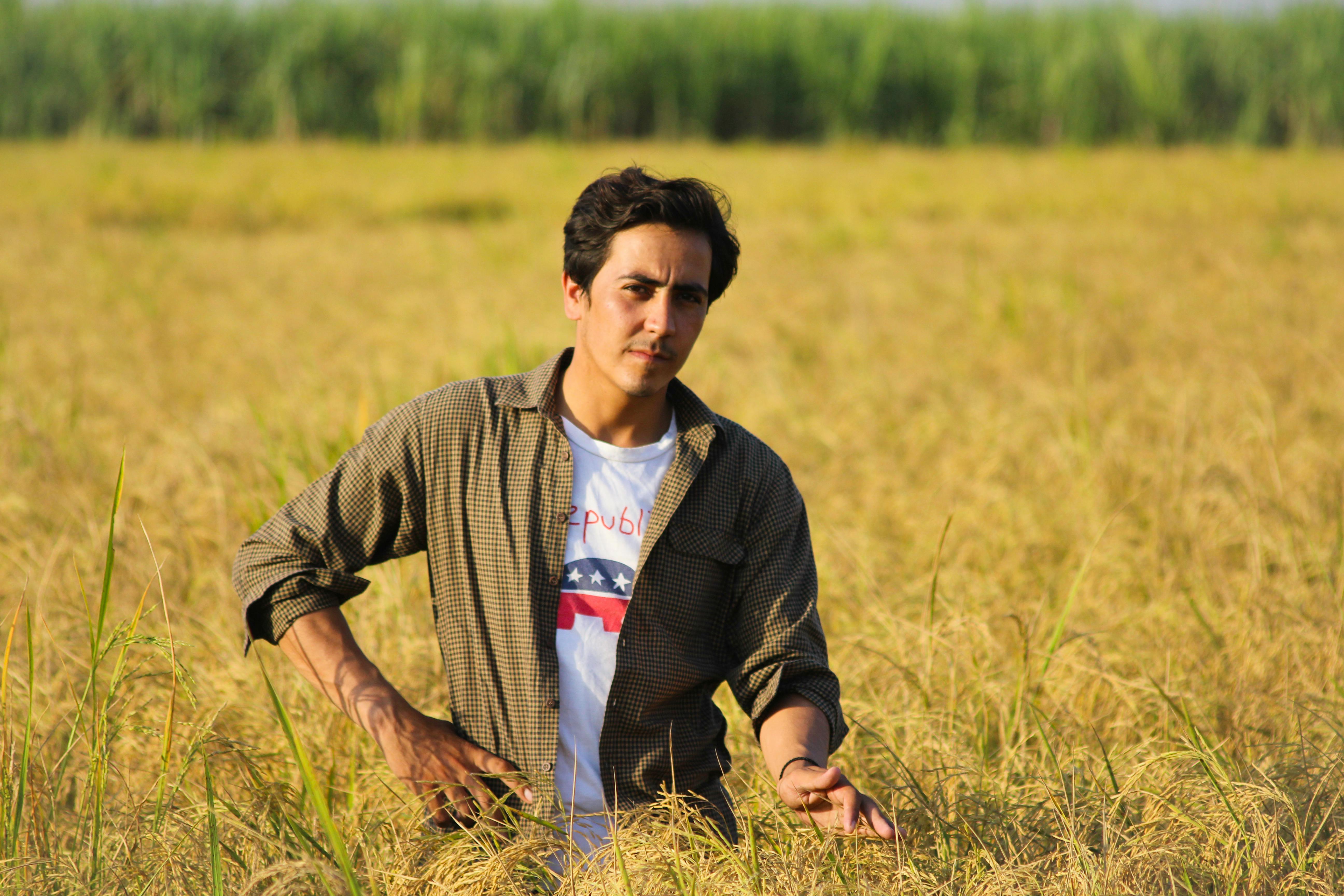 A young man stands confidently in a verdant wheat field, embodying rural charm and modern fashion.