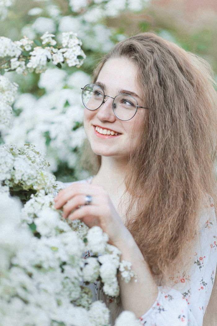 Young woman in round glasses smiling beside a hedge of white spring blossom — photo by Alina Rossoshanska on Pexels