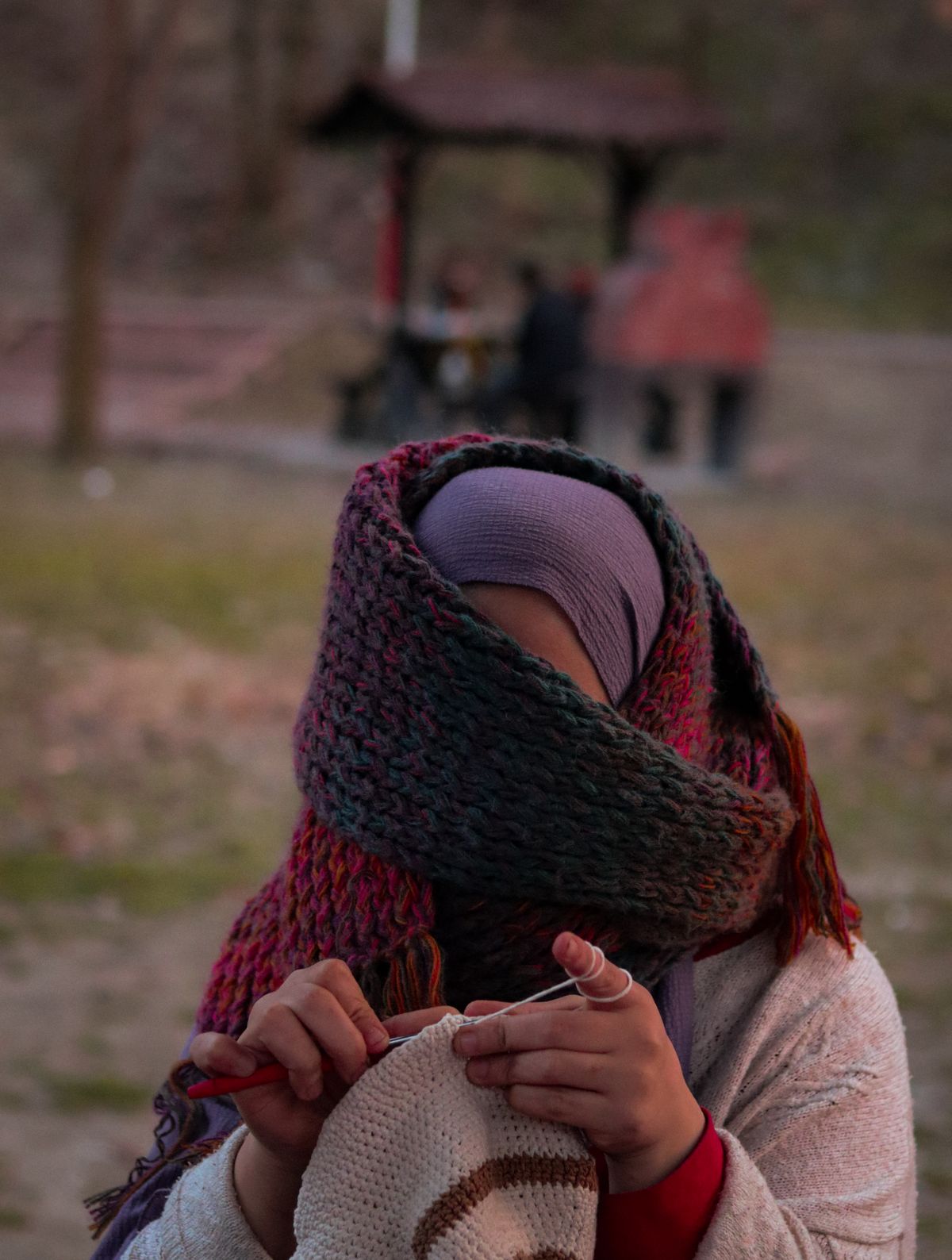 Woman wearing a warm scarf crocheting outside, creating handmade art surrounded by autumn scenery.