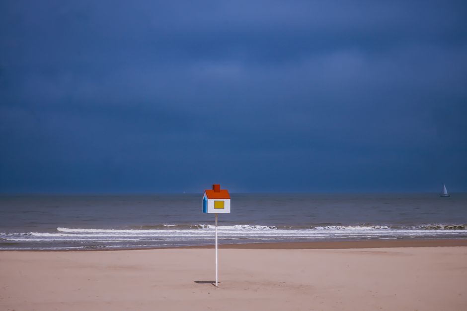 A small, colorful birdhouse stands alone on a calm and empty beach with a moody sky above.