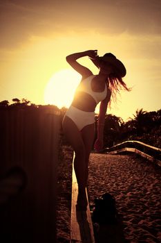 A woman in a bikini poses elegantly on a beach path during sunset, exuding summer vibes.
