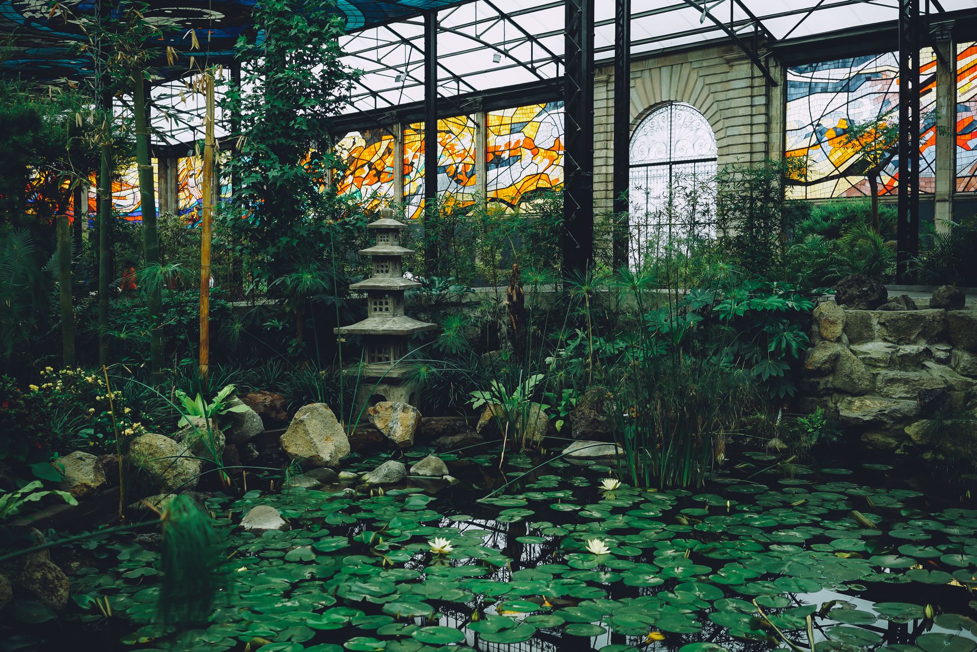 Lush greenhouse interior featuring a serene pond with water lilies and vibrant stained glass windows.