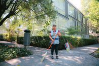 A woman casually sweeping a park walkway in Mexico City, surrounded by greenery and modern buildings.