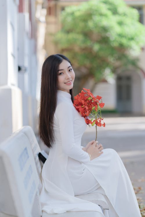 Young Brunette in a White Dress Sitting on a Bench and Holding Flowers