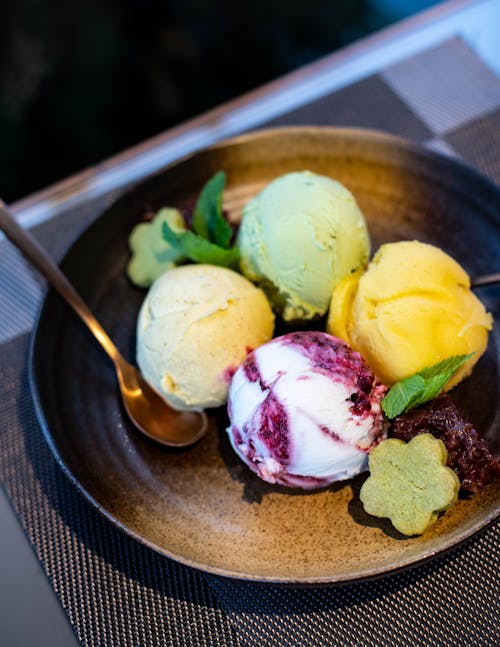 Scoops of ice cream assorted flavors with mint leaves and biscuits on modern plate with spoon placed on gray table mat