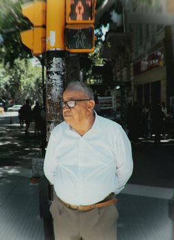 Senior man wearing glasses stands near a crosswalk in a sunny city street.
