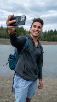 A cheerful man with a backpack capturing a selfie outdoors by a tranquil lake.