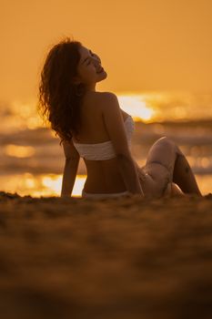 Woman in swimwear relaxes on a sandy beach at sunset, enjoying a peaceful moment by the sea.
