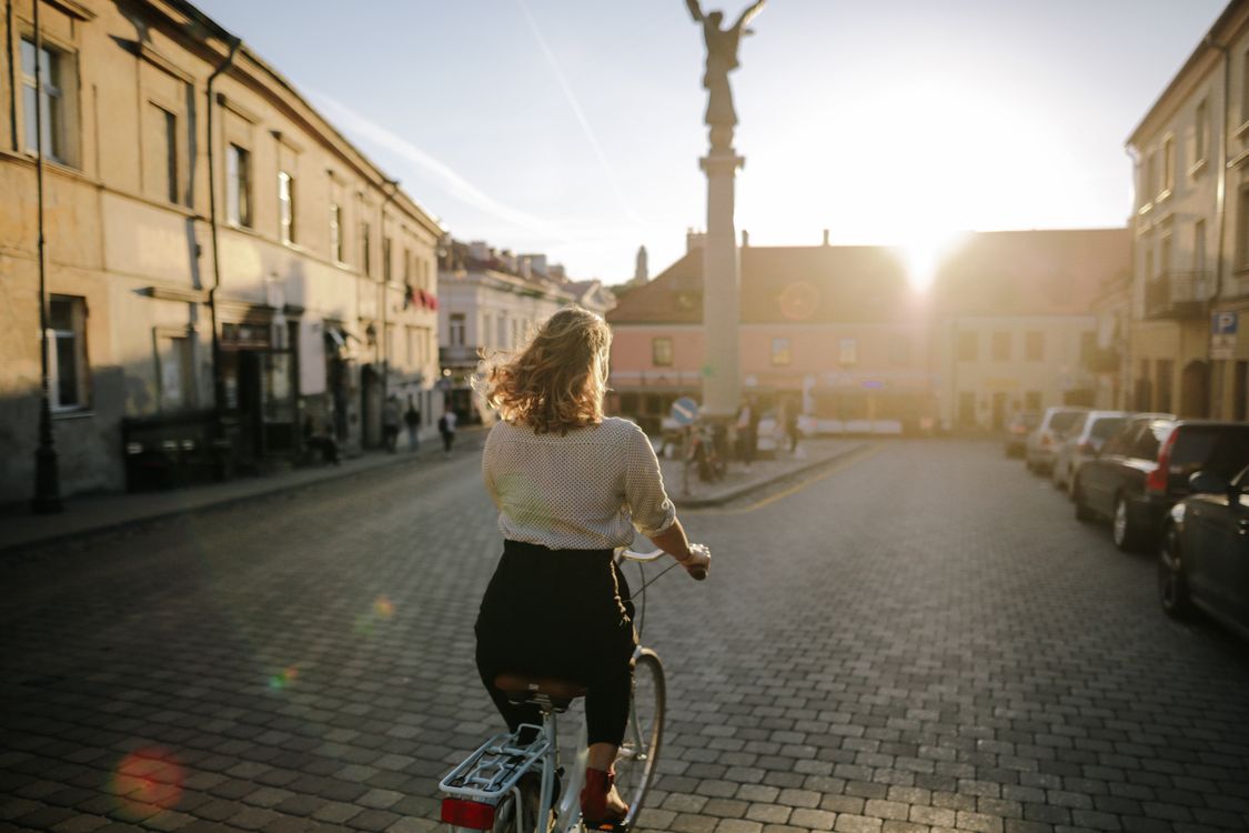 Free A woman rides a bicycle down a sunny, cobblestone street in a European city, creating a serene and joyful atmosphere. Stock Photo