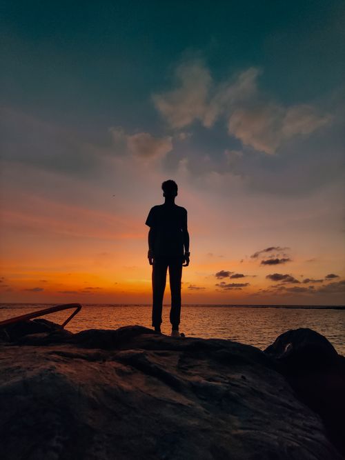 Free Silhouette of a person standing on rocks at the beach with a vibrant sunset sky in the background. Stock Photo