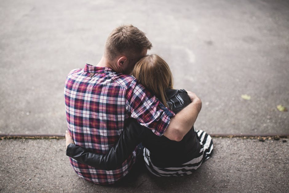 Man in Red White and Blue Check Long Sleeve Shirt Beside Woman in Black and White Stripes Shirt Hugging Each Other While Sitting on a Concrete Surface
